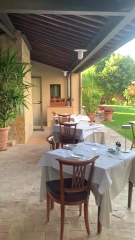 This is a video of an outdoor dining area with tables set for a meal. The tables are covered with white tablecloths, and chairs are placed around them. In the background, there is a lush green lawn with trees and rose bushes. There are also some potted plants and a gazebo with tables set for dining. The sun is shining, and the scene appears to be peaceful and serene.