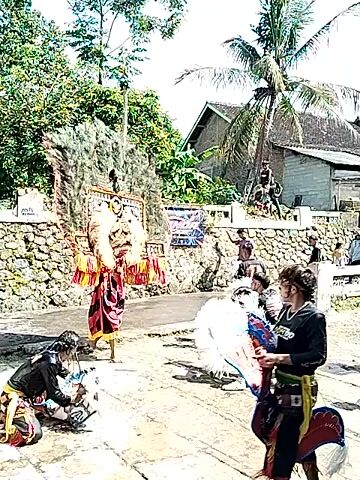 The video captures a traditional Indonesian dance performance. A man wearing a large, ornate mask and colorful costume dances to the rhythm of music. Several people in similar attire are also seen dancing. The performance takes place outdoors on a concrete surface, with a stone wall, trees, and buildings visible in the background. A coconut palm tree stands prominently on the left side of the frame. Several spectators can be seen watching the performance.