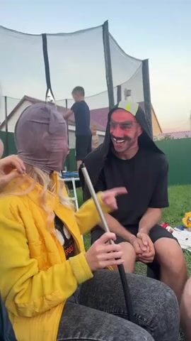 A person with their face obscured by a pinkish-purple mask, wearing a yellow jacket and grey pants, holds a metal rod. They are interacting with another person wearing a red devil mask and a black hooded sweatshirt. The devil mask prominently features red skin, exaggerated eyebrows, and a wide smile revealing uneven teeth. The scene takes place outdoors in a grassy area with a trampoline visible in the background. Two children are also present, one standing on the trampoline and another to the left of the frame. The lighting suggests it is dusk.