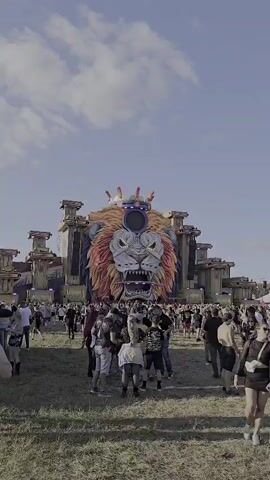 The video shows a large, elaborate stage setup at an outdoor event. The focal point of the stage is a massive, detailed sculpture of a lion's head wearing a crown. The lion's face is intricately designed with sharp teeth, glowing eyes, and a vibrant mane of orange and yellow. Below the lion's head is a platform where a DJ is visible performing. The stage structure appears to be made of concrete or stone-like material, with smaller structures flanking the lion's head. In front of the stage, a large crowd of people are gathered, many with their hands raised, suggesting they are enjoying a performance. The ground is covered in dry grass, and the sky is visible above, with some clouds. The overall impression is of a lively festival or concert.