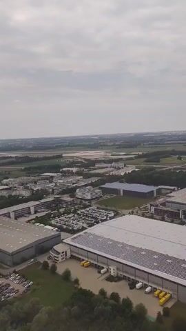 An aerial view of an industrial area. In the foreground, there are large warehouses with cars parked outside. In the mid-ground, there are more buildings, some of which appear to be office buildings, and more parking lots. In the background, there is a flat, rural landscape with fields and trees. The sky is overcast.