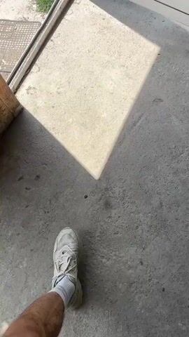 The video starts with a shot of a square of sunlight on a concrete floor just inside a doorway. The video then cuts to an outdoor scene. A shirtless man with a beard, wearing a white bucket hat, sunglasses, black pants, and white sneakers, is walking down a dirt path in a grassy field. He has a black backpack on. A woman with long, curly hair is walking ahead of him. The background shows green trees, a blue sky with puffy white clouds, and distant mountains.