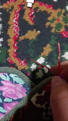 The video shows a person holding two red wires with exposed ends. When the ends of the wires are brought together, they create a small spark. The wires are held above a floral patterned cloth with green trim. A white USB connector is visible in the background on a multicolored rug. The person's hand is partially visible holding the wires.