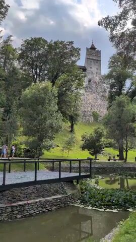 The video shows a scenic view of Bran Castle, a medieval fortress in Romania. The castle is perched on a hill overlooking a lush green landscape. In the foreground, a small pond with lily pads is visible, with a wooden bridge crossing over it. Several people are seen walking across the bridge and in the park area around the pond. The sky is cloudy, suggesting it might be a summer day. The overall impression is one of a tourist destination with historical significance and natural beauty.