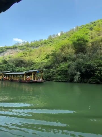 The video shows a view from inside a covered boat on a river. The water is a light green color, and there are visible wakes trailing behind the boat. On the right, another similar boat with a covered structure passes by. The shoreline features a steep hillside covered in lush green trees and vegetation. The sky is bright blue with a few scattered white clouds. People are visible inside the boat from where the video is being recorded.