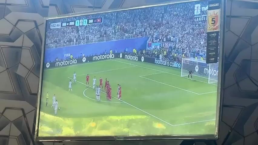 This is a wide shot video of a soccer game being played on a large television screen. The screen displays a soccer field with players in red and white uniforms. A penalty kick is about to be taken. A player in a red uniform is standing over the ball, and several players in white uniforms are forming a wall. The game clock shows 102:48. The background of the television screen shows a stadium filled with spectators wearing white shirts. The television is placed on a surface with a patterned wallpaper behind it. The scene is lit by ambient lighting.