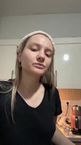 A woman with light blonde hair in a ponytail is shown from the chest up, facing the camera. She is wearing a black V-neck t-shirt and a cream-colored headband. She has a nose ring and gold hoop earrings. She is in a kitchen with white cabinets and a backsplash of small tan tiles. The woman holds two small green cucumbers in her hands. She bites one and then rubs one of the cucumbers on her forehead, cheeks, and chin. There are framed photos visible behind her, on top of the cabinets.