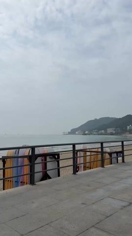 The video pans across a beach on a cloudy day. The camera is positioned on a raised walkway, overlooking the sandy beach and the ocean. To the right, there is a metal railing, and behind it, a rack filled with colorful surfboards stands. The beach itself is mostly deserted, with a few people walking along the shoreline and sitting on the sand. In the distance, a small island covered with trees and featuring a white building is visible. A sign in Korean is attached to the railing. The sky is overcast, creating a soft, diffused light across the scene.