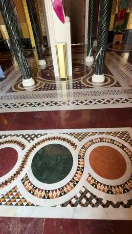 The video shows the interior of an ornate church. The camera starts by focusing on the intricately designed floor with geometric patterns and circular motifs in various colors. As the camera pans upwards, the viewer sees a series of spiraling black columns with gold accents supporting arches and alcoves. The walls are adorned with religious murals and paintings in gold and vibrant colors. The ceiling features a large, circular depiction of Jesus Christ surrounded by other figures and symbols. The church appears to be empty, with no people visible. The atmosphere is calm and reverent, emphasizing the architectural beauty and religious art.