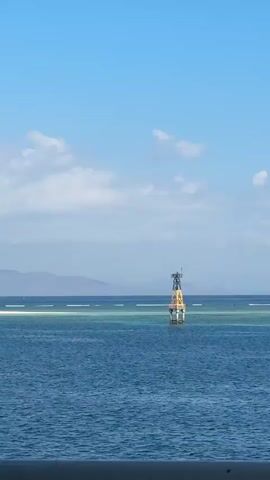 The video displays a wide shot of a calm, blue ocean under a clear sky with scattered white clouds. A buoy, primarily yellow and white with a black top, stands in the center of the frame. It appears to be a navigational marker. In the distance, a faint, hazy shoreline with mountains is visible. The water in the foreground is a deep blue, transitioning to lighter turquoise hues near the buoy, suggesting shallow water or a reef. The horizon is well-defined, separating the sea from the sky. A dark railing or structure is partially visible at the very bottom of the frame, indicating the viewpoint is from a vessel or elevated platform. The overall scene is serene and depicts a clear day at sea.
