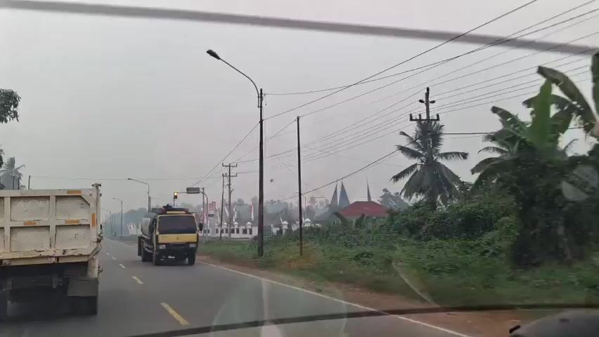 The video shows a view from a vehicle traveling on a road. On the left, a white dump truck is visible. In the center of the road, a yellow tanker truck drives past. On the right side of the road, there is dense greenery and trees, including palm trees. Several utility poles with power lines stretch across the scene. In the distance, a line of white fences with flags and what appear to be buildings or monuments is visible. The sky is overcast and hazy.
