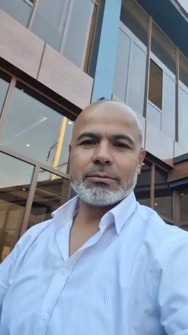 A man with a shaved head and a gray beard is looking at the camera and smiling. He is wearing a white collared shirt. He is standing in front of a modern building with large windows. The building appears to be made of wood and glass. There are trees and plants visible in the background. The sky is blue.
