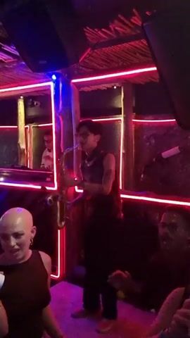 The camera films a nightclub, its patrons, and a DJ in a booth. A musician plays the saxophone, while patrons hold cups of drinks and bob and dance to the music. Neon lights are on the walls and ceiling. The DJ is in a small booth, looking out into the crowd. A bald woman stands near the camera, holding a cup and smiling. The ceiling has a woven texture. Speakers are visible in the upper corners.