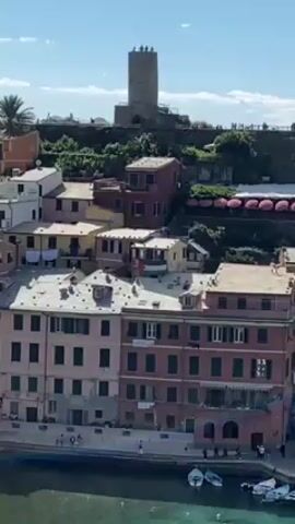 The video reveals a picturesque coastal town nestled on a rocky promontory, overlooking a small harbor dotted with boats. Buildings are painted in vibrant, varied hues, a tower stands atop the promontory, with a few people visible at its peak. Clear blue sky with a few fluffy clouds. The camera zooms out gradually.