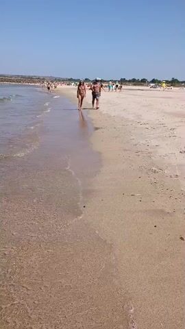 Two individuals, one male wearing shorts and a hat, and one female wearing a bikini, walk along the wet sand of a beach near the water's edge. The tide is low, and small waves are washing onto the shore. In the background, other beachgoers are visible, some in the water and others on the sand. The sky is clear and blue, and the sun is shining. The overall environment appears to be a typical sunny day at a crowded beach.