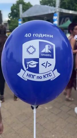 A close-up shot of a blue balloon with a crest and text in Bulgarian. The crest features a graduation cap, a microchip, and code brackets. The text at the top reads "40 ГОДИНИ" (40 YEARS), and at the bottom reads "НПГ по КТС" (NPG of KTS). The background is blurred but shows a stage with a band playing, decorated with blue and white balloons, and an audience gathered. Several people are visible, some facing the stage and others turned away from the camera. The overall scene appears to be an outdoor event, possibly a celebration or anniversary.