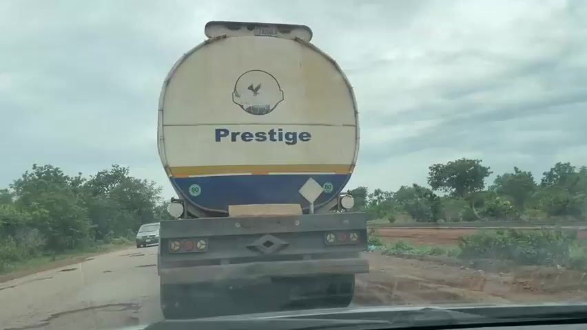 The video shows the view from inside a vehicle driving on a paved road with a tanker truck labeled "Prestige" in front. The sky is overcast with clouds. To the left of the road is a line of green trees and bushes. To the right, there is a dirt road and more vegetation. There is another vehicle, a silver minivan, visible to the left of the tanker truck. The road appears to be in poor condition with visible cracks and potholes. The tanker truck is white with blue and yellow accents and a circular logo above the word "Prestige". The rear of the truck has multiple red lights and reflectors. The video appears to be taken during daylight hours.