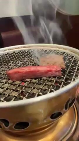 In a close-up shot, slices of raw red meat are placed onto a small circular grill with a gold-colored base. The grill sits on a table and is heated from below, causing smoke to rise from the cooking meat. One piece of meat appears to have been cooking for longer as it has browned more. The person uses a pair of tongs to flip a piece of meat onto the grill. The background is out of focus and appears to be a restaurant setting with dark wooden furniture and white walls.