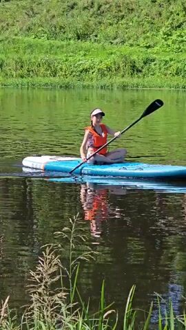 A person wearing an orange life vest and a pink hat is sitting cross-legged on a blue paddleboard in the middle of a calm river. The person is holding a paddle with both hands. The water reflects the person and the paddleboard. Green foliage lines the riverbank in the background. There are tall reeds growing out of the water in the foreground.