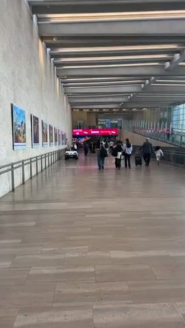 The video shows a wide, well-lit hallway in what appears to be a modern airport or train station. The hallway has a high ceiling with a grid of lights and a long row of windows on the right side, offering a view of buildings and the sky. The floor is made of large, light-colored tiles.

On the left wall, there are several framed pictures or posters displayed at intervals. A few people are walking in the hallway. Towards the center, a small white electric vehicle with a driver is visible. Further ahead, a large digital screen displays the time "07:28" in bright pink digits.

On the right side of the hallway, a moving walkway or travelator is in operation, with several people walking on it, some pulling luggage. The travelator has a dark-colored base with some text visible, including "DREAMER" and "Life Chan".

The general atmosphere is one of transit and movement, with people going about their journeys. The lighting is bright and even, creating a clean and functional environment. The overall impression is of a busy, modern transportation hub.