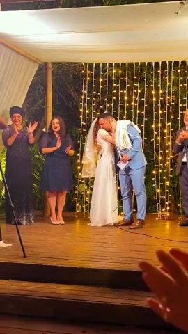 A wedding ceremony takes place under a canopy. A bride in a white dress and veil stands with a groom in a blue suit. The groom is kissing the bride. Two women stand to the left, clapping. A man in a suit stands to the right, clapping. A man with a microphone walks across the foreground. The background is decorated with string lights.