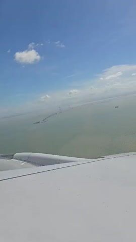 The video displays a wide-angle aerial view from an airplane window. The foreground is dominated by the wing of the aircraft, which is white and appears clean. Beyond the wing, a vast expanse of greenish-blue water stretches to the horizon. In the distance, a long bridge or causeway is visible, stretching across the water, with several structures or supports appearing along its length. A few ships or boats are also discernible on the water. The sky above is a vibrant blue, dotted with scattered white clouds, some of which are illuminated by the sun. The sun itself is visible as a bright glare near the horizon on the right side of the frame. The overall impression is one of flight over a body of water with a significant man-made structure crossing it, under a partly cloudy sky.