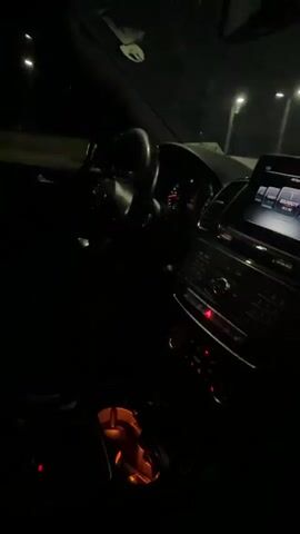 A nighttime video from the interior of a car traveling on a highway lined with streetlights. The dashboard, steering wheel, and central console are visible in the dimly lit interior. A digital display on the dashboard shows radio information. The view through the windshield reveals oncoming and passing vehicles on the multi-lane highway.