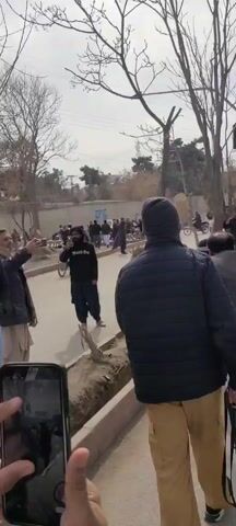 The video shows a street scene with a group of people, some of whom appear to be armed. Several men are wearing traditional clothing, while others are in jackets or camouflage. One man is carrying a rifle. The group is walking down the street, and some are pointing or gesturing. The environment appears to be a residential area, with buildings and trees visible in the background. The weather seems mild, and the sky is overcast.