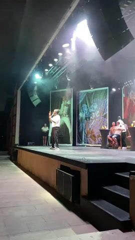 A person stands on a stage in front of several large portraits. The person is holding a microphone and appears to be speaking or performing. There are also other people seated in the background. The stage is lit and there is a spotlight shining down. The environment appears to be an indoor performance venue.