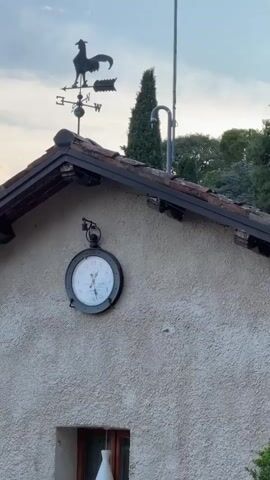 A video shows the exterior of a building. In the foreground, a clock is mounted on a rough-textured wall. Above the clock, a roof with dark tiles is visible, and on the peak of the roof, a black rooster-shaped weather vane is mounted. Behind the building, trees are visible, and the sky is cloudy. The video pans from left to right, revealing more of the roofline and the sky. The clock shows the time to be approximately 8:15.