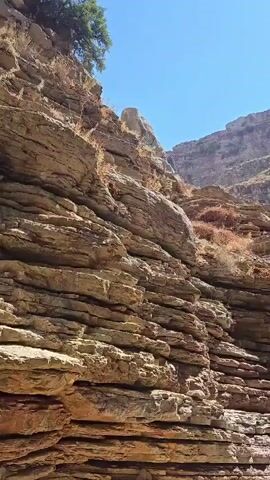 The video shows a narrow canyon with layered rock formations. Two people are present. One person, a male wearing a white t-shirt and black shorts, is climbing up the rocky wall of the canyon. Another person, a female wearing a white dress, stands on the canyon floor observing. The canyon floor is sandy and rocky. The sky is visible at the top of the canyon, appearing clear and blue.