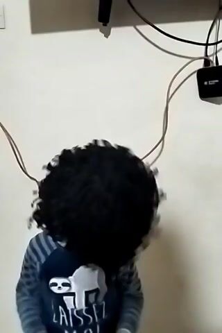 A child wearing a large, dark, curly wig stands in front of a light beige wall under a TV. The child is wearing a striped long-sleeved shirt with a sloth design. The TV is showing an animated image of a man in traditional clothing playing a guitar. Various cables and a small black box are visible on the wall between the TV and the child. The child appears to be singing and laughing.