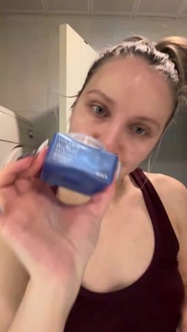 A woman is shown applying a blue cream from a jar to her face with a cotton pad. She is fair-skinned, with blonde hair pulled back in a bun, and wearing a maroon tank top. The background shows a white bathroom with a washing machine and a door visible. She first holds up the jar of cream, and then proceeds to apply the cream to her forehead, cheeks, and chin using a cotton pad in her right hand.
