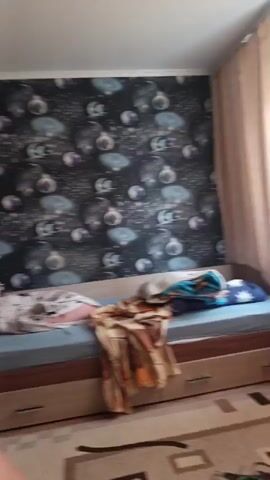 This video captures a tour of an indoor living space, likely a bedroom and adjoining areas, in Sukhodol, Russia. The initial frame shows a close-up of a bed with a person lying down, partially covered by a patterned duvet. The wallpaper behind the bed features a celestial theme with planets and stars.

The camera then pans to reveal more of the room, including a dresser with drawers and a television on top. Further along, a large wardrobe stands against the wall, and shelves are visible, holding various items. A patterned rug covers the floor. The next scene shifts to a desk area, where a laptop and a computer mouse are placed. A doorway in the background leads into a kitchen area, which appears to be modern and well-lit, with white cabinets and countertops.

The overall impression is that of a lived-in home. The lighting suggests it could be daytime, as there is no indication of artificial lighting being the primary source of illumination, and the overall brightness is consistent with daylight. There are no specific weather cues as the scene is entirely indoors. The presence of personal belongings and furniture indicates a residential setting. No specific emotions are conveyed, beyond a sense of casual observation or a private walkthrough. There is no visible text in the video.