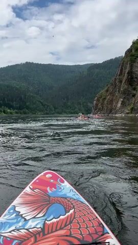 A first-person perspective video shows a person on a paddleboard with a koi fish design on the front. The paddleboard is on a wide river with choppy water. In the distance, several other people on rafts are visible, also on the river. The riverbanks are steep and covered in lush green trees. The sky is overcast with some patches of blue.