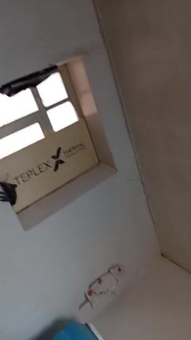 The video begins with a blurry shot of a white ceiling with a few dark specks. It then pans to the right, revealing a window with sunlight streaming through. Below the window, there are piles of clothes. The camera then moves to show a standing fan with orange blades, and a white electrical outlet. The fan is positioned on the right side of the frame, and the outlet is on the wall behind it. The room appears to be a bedroom, with a large pink blanket visible on what looks like a bed.