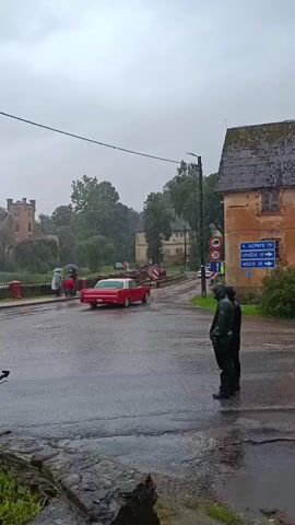 A person wearing a dark green raincoat and helmet stands on the right side of the frame, facing left. The person is in the foreground and slightly to the right. Behind the person is a tan building with a dark roof. To the left, a red car drives away from the viewer down a wet road. In the background, a castle-like building with a red roof is visible on the left, with trees and other buildings in between. People holding umbrellas are walking on the left side of the road. The ground is wet, indicating it is raining. A street sign with arrows pointing to "AUPLĪDE 19", "UPSIDE 19", and "MĒŽI 23" is visible on the tan building. Traffic signs are also present near the road.