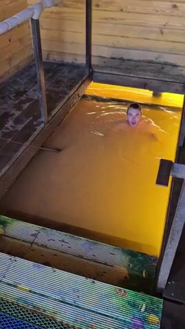 A person is in a yellow body of water, possibly a pool or a bath, within a wooden structure. The person's head and upper chest are visible above the water. The water is rippling, indicating movement. There are metal railings and wooden planks surrounding the water. The lighting appears to be coming from beneath the water, giving it a yellow glow.