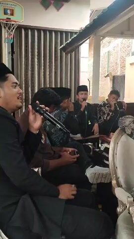 The video shows a group of men gathered outdoors, possibly under a covered patio. Several men are holding microphones and appear to be singing or speaking. One man in the foreground on the left is holding a microphone and wearing a black shirt. To his right, another man in a patterned shirt is also holding a microphone. Further back, a man in a black shirt and cap is speaking into a microphone. Another man in a patterned shirt sits beside him, also holding a microphone. The environment appears to be a residential area with buildings visible in the background. The men are seated, and the atmosphere suggests a musical performance or a gathering.