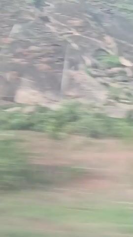 The video shows a landscape with a steep, rocky hillside alongside a field, shot from a moving vehicle. The hillside is mostly gray and brown with sparse vegetation. The field appears to be divided into sections of green grass and brown soil. The camera moves quickly, blurring the foreground and providing a passing view of the landscape. At one point, a tree is briefly visible against the background of the hillside. The sky is overcast, casting a flat light over the scene.