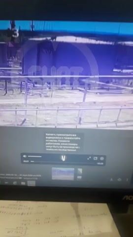 The video shows a computer monitor displaying a video of a large cylindrical object engulfed in flames, possibly a tank or industrial container. The video has a watermark across it. Beneath the screen, there is a piece of paper with handwritten notes and calculations. The monitor itself is labeled "AOC". The video footage seems to show a fire or explosion incident at an industrial site.