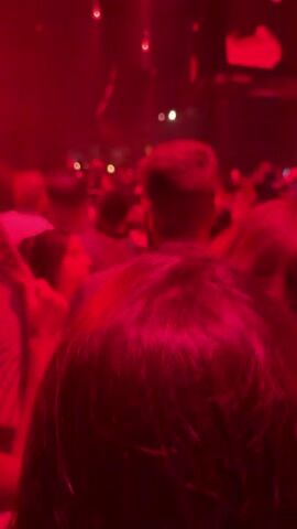 A dimly lit, crowded indoor venue, likely a club or concert hall, is illuminated by vibrant red lighting and occasional white spotlights. The camera, held at eye level, captures the backs of several people in the foreground, their forms blurred by the low light and movement. Beyond them, a dense crowd of individuals can be seen, their faces obscured. Some appear to be dancing or conversing, while others are more static. The atmosphere suggests a lively event with music playing, though no sound is present in the visual. In the background, structural elements of the venue are visible, including what appear to be speakers hanging from the ceiling and architectural details that hint at a somewhat rustic or industrial design. The overall impression is one of energetic, albeit dimly lit, social gathering.