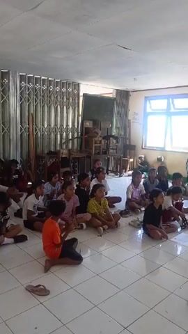 A group of children are sitting on the floor of a room facing towards the camera. They appear to be in a classroom setting. There are windows in the background and a door with a metal gate. Some of the children are looking at the camera, while others are looking down at papers on the floor. There are papers scattered on the floor in front of some of the children. One child in the foreground is wearing an orange shirt and black shorts and is holding something in their hands. Another child to their left is wearing a striped shirt. A person, possibly a teacher, is standing on the right side of the frame, facing away from the camera. The room has tiled flooring and a light-colored wall. There is a wooden table and chairs in the background. The overall lighting suggests it is daytime.