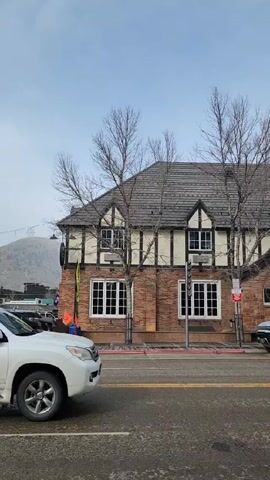 The video shows a street in front of "The Gant Aspen" hotel. Cars are parked along the street in front of the hotel. The building is a large two-story Tudor-style structure with a brick facade and a dark gray tile roof. Several bare trees stand in front of the building. The sky is overcast. The video pans from right to left, showing more of the hotel and parked cars.