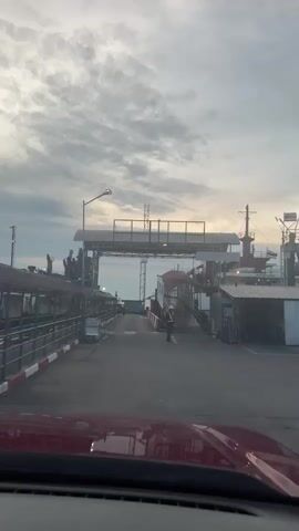 The video shows a car driving towards a ferry terminal. The time of day appears to be early morning or late afternoon, as indicated by the soft, diffused light and the cloudy sky. The weather is overcast. The scene is a paved road leading to a ferry. On either side of the road are covered structures with railings and what appear to be ticket booths or waiting areas. A ferry is docked at the terminal, with a ramp extended for vehicles to board. A person wearing a high-visibility vest is standing near the ramp, possibly directing traffic or assisting passengers. The car's dashboard is visible in the foreground, showing a speed of 10. The overall atmosphere is one of routine travel and transport. The location is identified as unknown, Thailand.