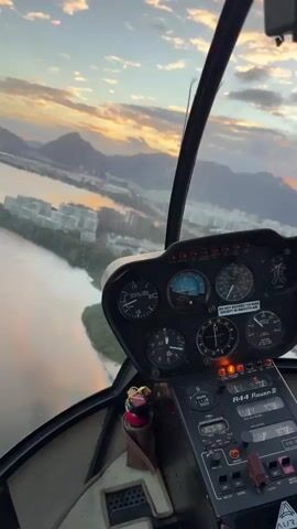 The video displays a first-person perspective from the cockpit of a helicopter, specifically an R-44 Raven II, as indicated on the visible control panel. The instrument panel, featuring multiple circular gauges, a radio stack, and various switches, occupies the right side and bottom-center of the frame. Through the helicopter's windscreen, a vast body of water is visible below and to the left, reflecting the ambient light. Beyond the water, a landmass with numerous urban buildings and green foliage stretches into the distance. In the far background, a range of mountains rises against the horizon. The sky is partially cloudy, illuminated by warm, soft light suggesting either sunrise or sunset, with hues of orange, pink, and blue. The helicopter appears to be in stable flight, with the external scenery slowly passing by.