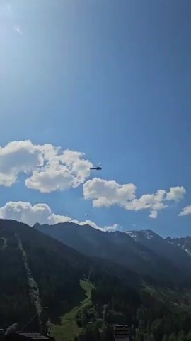 The media file shows a helicopter flying over a mountainous landscape. The helicopter appears to be on a mission, possibly for rescue or transport, as it is carrying a sling load. The scene is set in a valley with steep, tree-covered slopes and snow-capped peaks in the background. The sky is clear blue with scattered white clouds, suggesting a sunny day. The time of day appears to be midday, given the bright sunlight. There are some buildings visible in the lower part of the frame, indicating a small settlement or village at the foot of the mountains. The overall impression is one of rugged natural beauty and human activity in a remote mountainous area. No visible text is present in the media file.