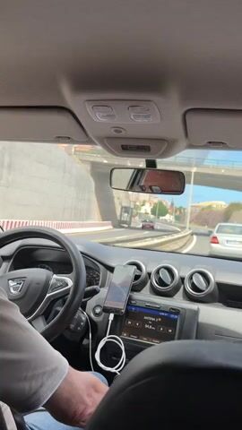 The view is from the back seat of a car, looking forward at the driver, dashboard, and the road ahead. The car is a Dacia, as seen on the steering wheel. The driver, wearing a white shirt and blue jeans, has his hands on the steering wheel. A smartphone is mounted on the dashboard, displaying a map, and is connected to the car's audio system with a white cable. The car radio is tuned to ANTENA 3, 94.8 FM. Outside, the car is driving on a highway lined with concrete barriers and rocky hillsides. Other cars are visible on the road. The weather appears sunny, and buildings can be seen on the hills in the distance. The car is approaching a tunnel.