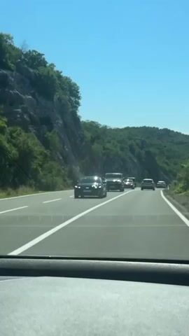 The video captures a convoy of black vehicles traveling on a multi-lane highway under a clear blue sky. The footage is taken from inside a moving car, looking forward through the windshield. The lead vehicle is a black sedan, possibly a Mercedes-Benz E-Class, featuring a red light bar across its front and the word "POLICIJA" (Police) visible below the grille. Immediately behind it is a black Mercedes-Benz V-Class van, followed by several other black SUVs and sedans, maintaining a tight formation. The highway is bordered by a steep, rocky, tree-covered hillside on the left, and more green, forested mountains in the distance on the right, suggesting a scenic, mountainous region. The weather appears sunny and clear. Background music is audible throughout the clip.