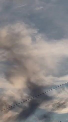 The video begins with a close-up of a dark, swirling cloud formation against a lighter sky. The camera then pans downwards to reveal a wide expanse of sky filled with various cloud types, ranging from dark and ominous to light and wispy. As the camera continues to descend, it captures a beautiful sunset with warm orange and yellow hues illuminating the clouds. In the foreground, silhouetted against the sunset, are the tops of structures that appear to be rooftops and fences. A single utility pole is also visible. The final shot focuses on the lower part of the scene, showing a dark, blurry foreground with what appears to be a bicycle and some debris. The overall impression is one of a dynamic sky with a dramatic sunset, viewed from a somewhat rural or less developed area.